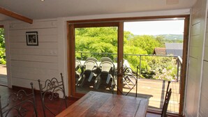 Un comedor con mesa de madera y sillas de metal, un balcón con barandilla y vistas a zonas verdes y colinas.
