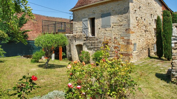 Una casa de piedra con techo de tejas rojas, un balcón pequeño y un jardín con rosales.