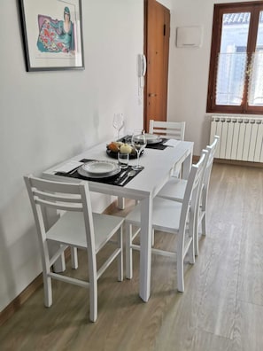 Un comedor con una mesa blanca para cuatro, sillas blancas, un cuadro en la pared, una puerta de madera y una ventana con cortinas de encaje.