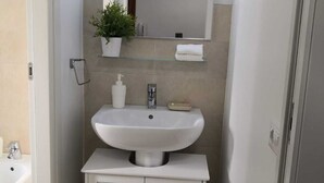 Um banheiro com pia branca, espelho acima, planta em vaso e armário abaixo.