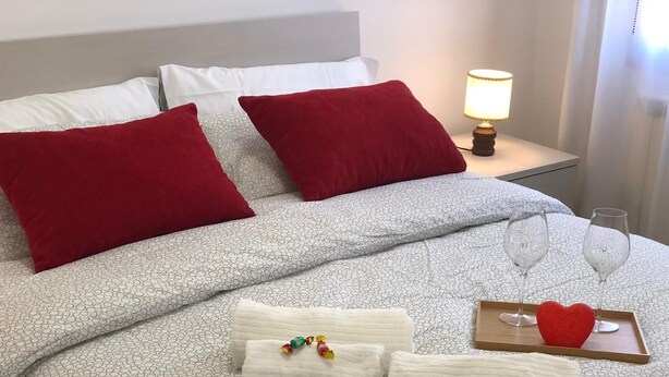 Cama bem arrumada com almofadas vermelhas e brancas, um tabuleiro com duas taças de vinho e um objeto em formato de coração vermelho, e várias toalhas brancas com balas coloridas.