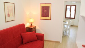 Sofá vermelho em um quarto com uma mesinha de cabeceira de madeira, um abajur e duas fotos emolduradas na parede. Há uma janela com vista para uma área de jantar com mesa e cadeiras.