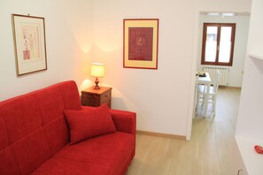 Ein rotes Sofa in einem Raum mit einem hölzernen Nachttisch, einer Lampe und zwei gerahmten Bildern an der Wand. Ein Fenster bietet Blick auf einen Essbereich mit Tisch und Stühlen.