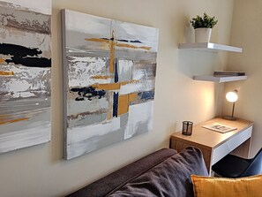 Un salon moderne avec des œuvres d’art abstraites, un bureau avec une lampe et une plante posée sur une étagère.
