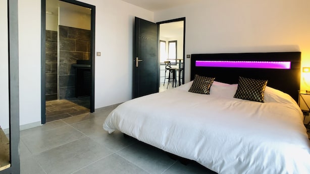 Un dormitorio moderno con una cama grande, cabecera con iluminación LED, baño independiente con puerta de vidrio y un área de comedor visible a través de una puerta abierta.