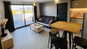 Una sala de estar moderna con un sofá, una mesa de centro y una mesa de comedor. Hay un ventanal corredero que da a un balcón con vistas a las colinas.