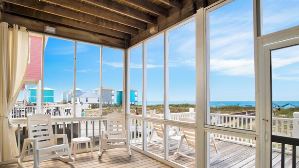 A wooden deck with white rocking chairs and a view of beach houses.