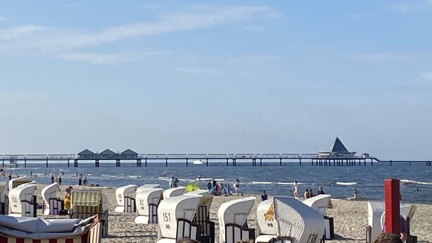 A beach with beach huts numbered 151, 152, and 153, a pier extending into the sea, and a building with a pointed roof.