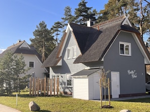 Two-story house with a steeply pitched roof and thatched roof extension, set amidst a well-maintained lawn and a wooden fence.