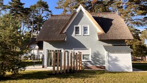 A two-story house with a grey exterior, a thatched roof, and a wooden fence in front.