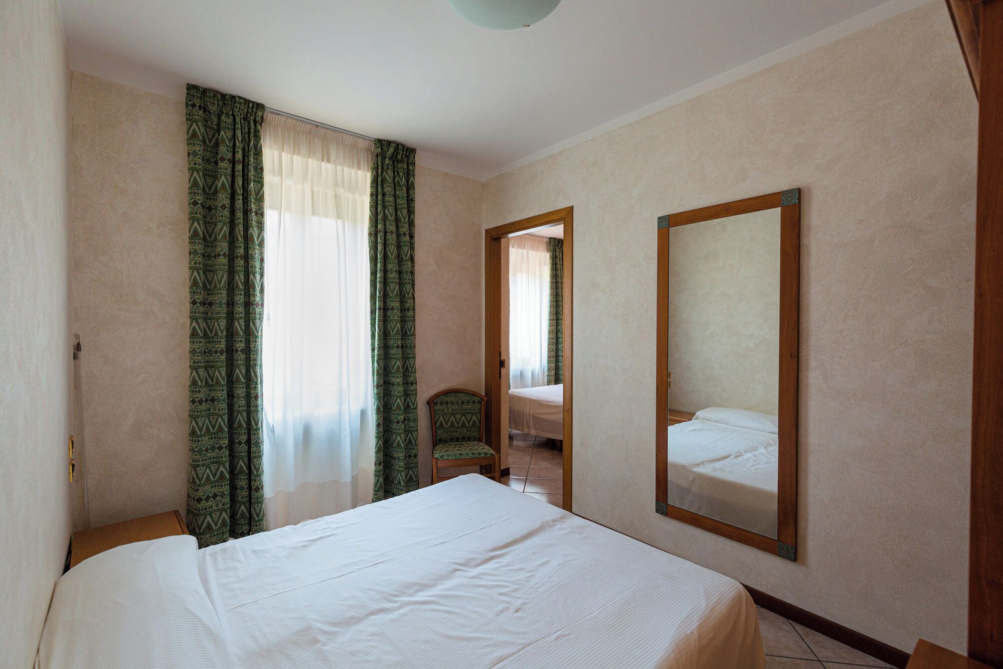 A hotel room with a large bed, a wooden chair, a mirror, and a door leading to another room.