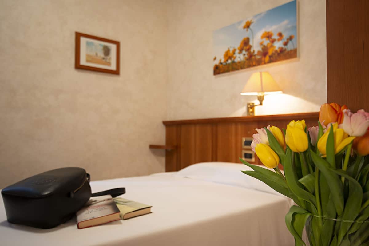 A hotel room with a bed, a bedside table with a lamp, a black bag, a book, and a bouquet of tulips.