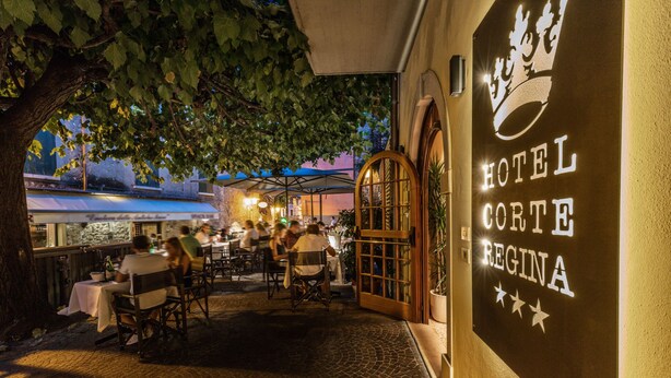 Un área de comedor al aire libre con mesas y sillas, un gran árbol que proporciona sombra, y un edificio con un letrero que dice "HOTEL CORTE REGINA".