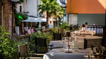 Un área de comedor al aire libre con mesas dispuestas para dos, rodeada de vegetación y elementos arquitectónicos.