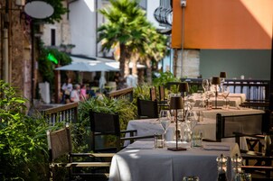 Un área de comedor al aire libre con mesas dispuestas para dos, rodeada de vegetación y elementos arquitectónicos.
