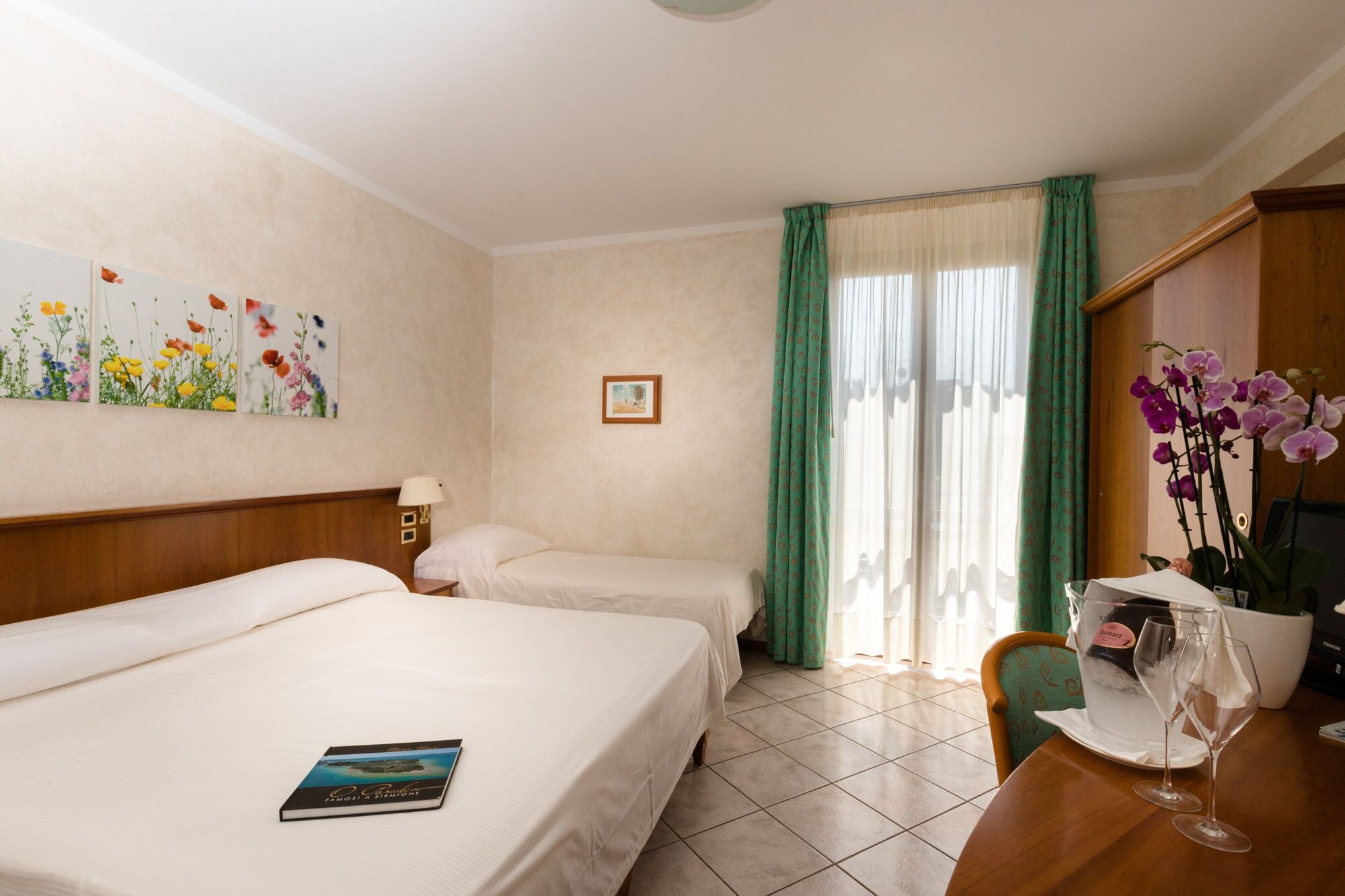 A hotel room with two beds, a dining table with a vase of flowers, and a window with green curtains.