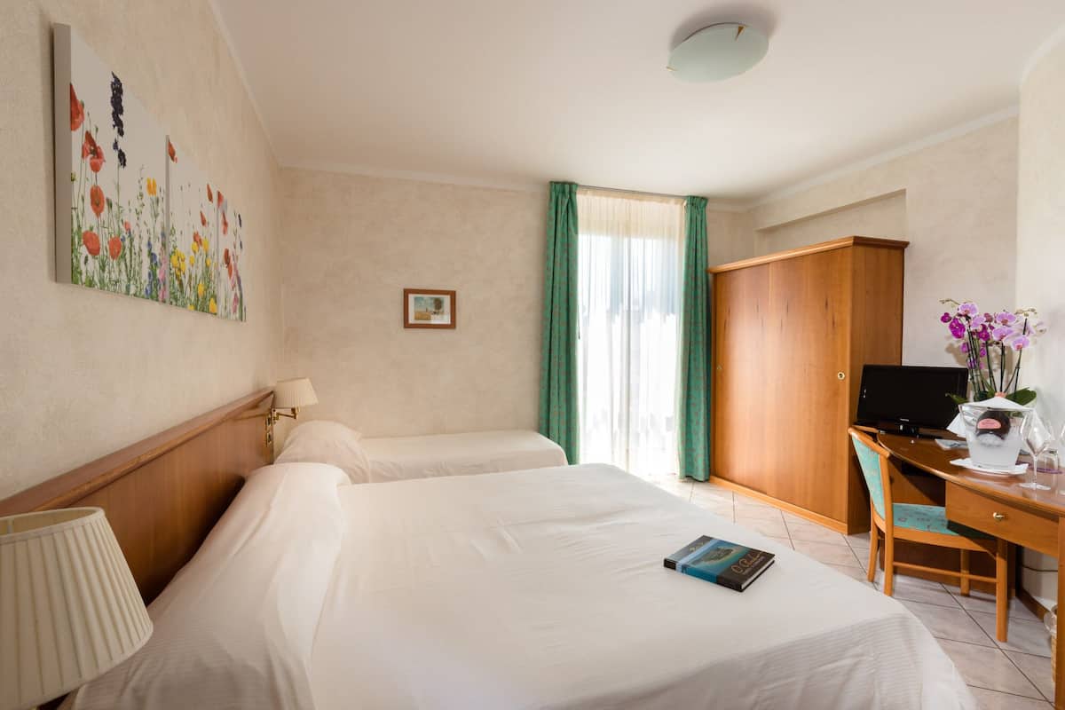 A hotel room with two beds, a desk with a computer, a lamp, a framed picture on the wall, and a window with curtains.