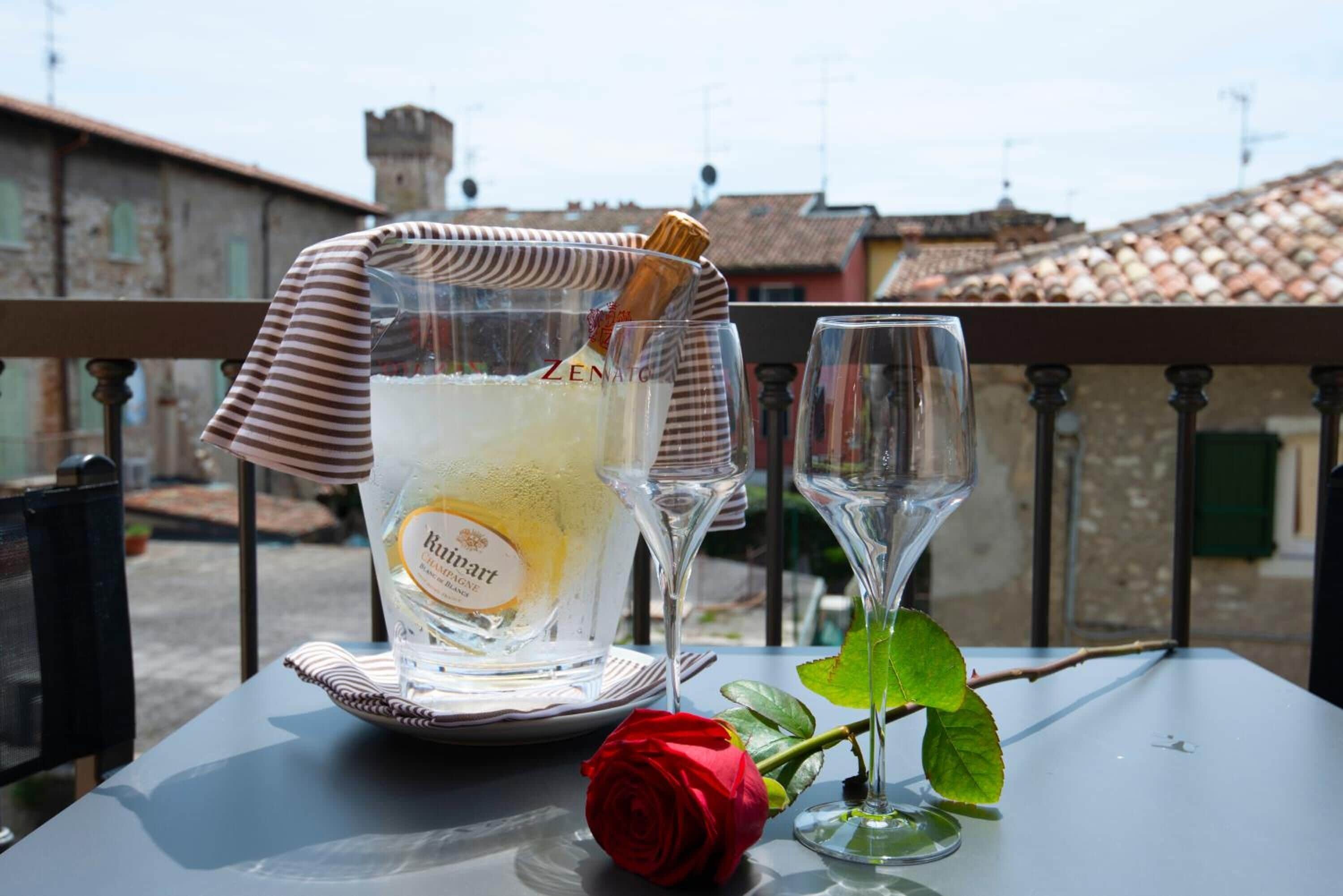 Ein Glas RUTINO Wein, ein Glas Champagner und eine rote Rose auf einem Tisch mit einer gestreiften Tischdecke. Dazu ein Glas Wasser.