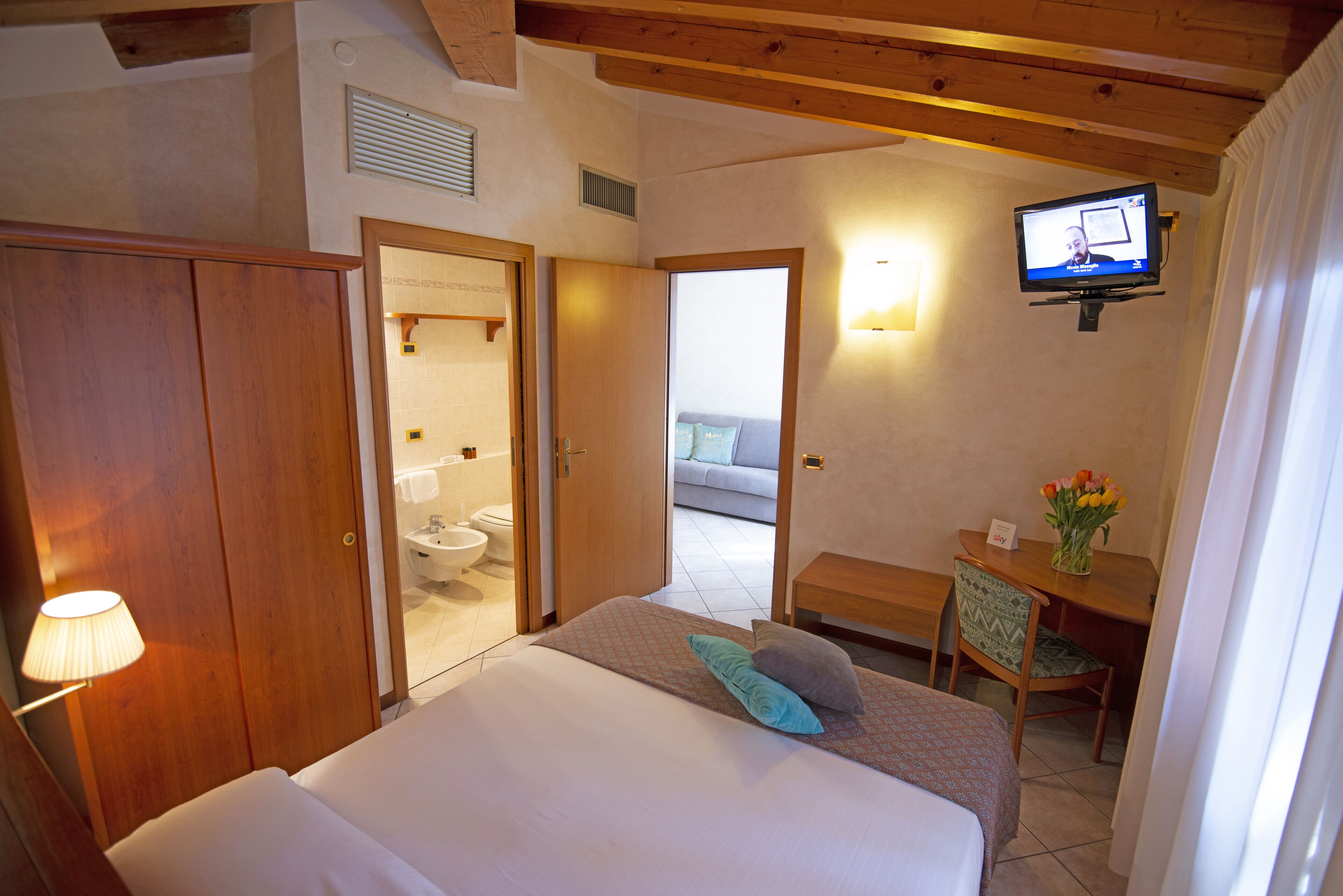 A hotel room with a bed, a desk with a chair, a television mounted on the wall, and a bathroom visible through an open door.