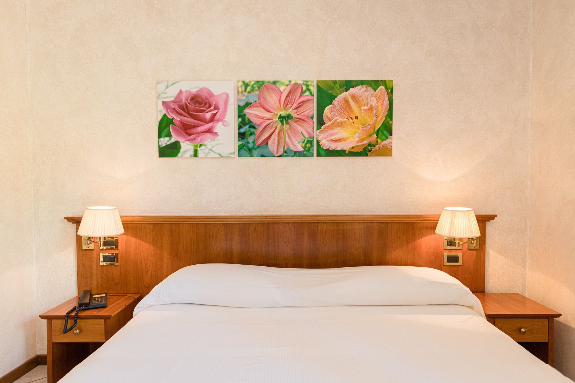 Habitación de hotel con cabecera de madera, cama con ropa blanca, dos mesitas de noche con lámparas y un cuadro con tres pinturas florales.