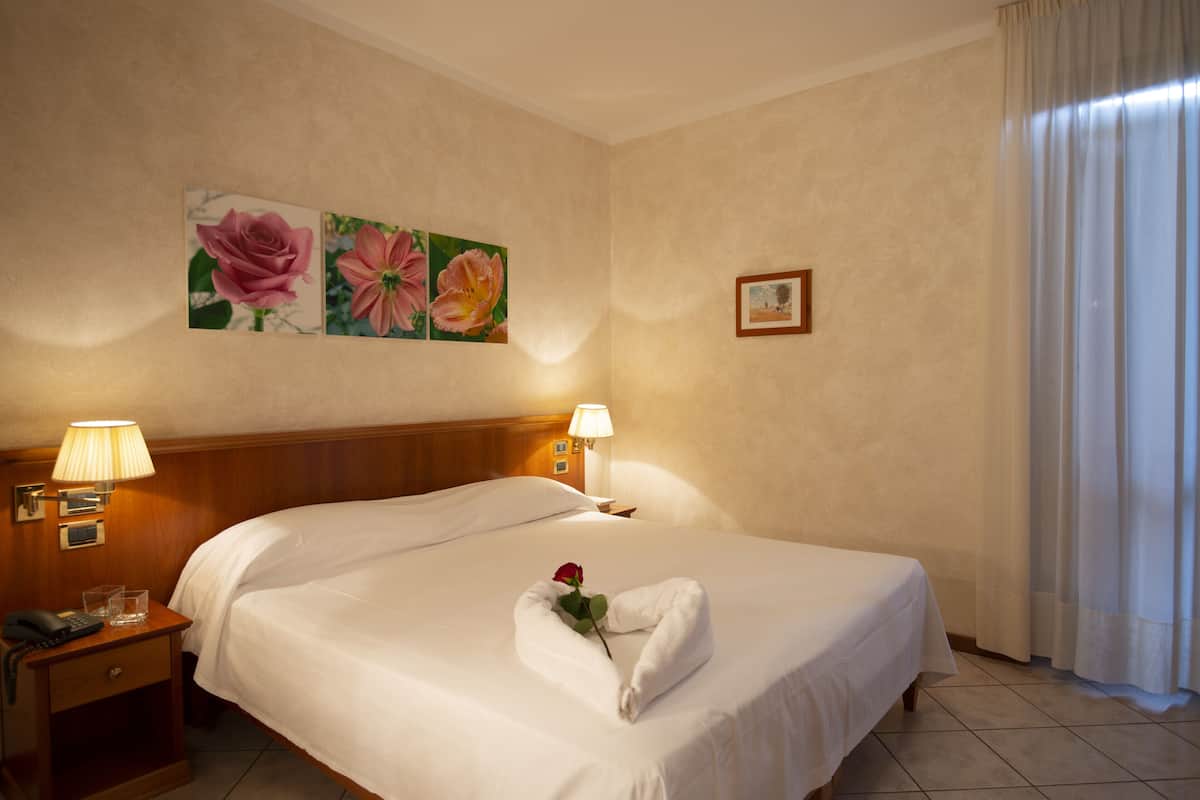 A hotel room with a large bed, two bedside lamps, a nightstand with a phone, a framed picture on the wall, and a window with curtains.