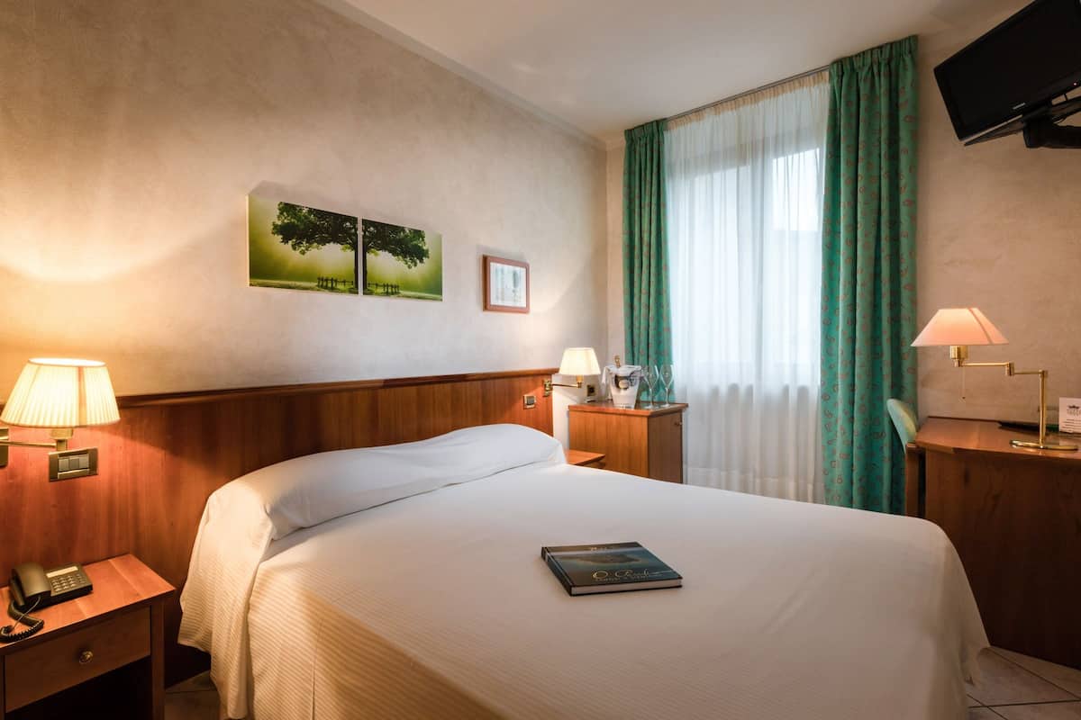 A hotel room with a bed, a nightstand with a telephone, a bedside table with a lamp, a picture on the wall, and a window with curtains.