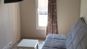 Une petite pièce, peu meublée, avec un canapé, une petite table, un téléviseur fixé au mur et une fenêtre avec des rideaux.