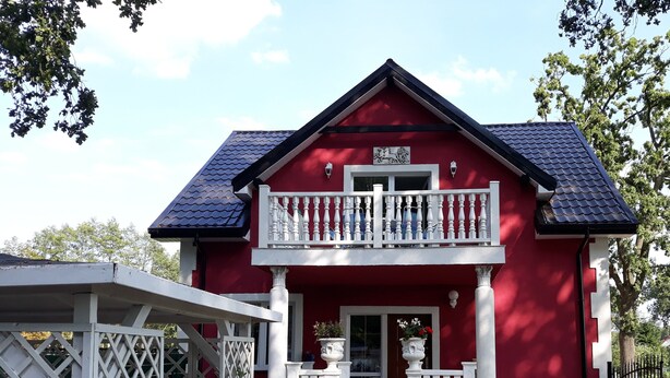 A red two-story house with a balcony, a white fence, and a pathway leading to the entrance.