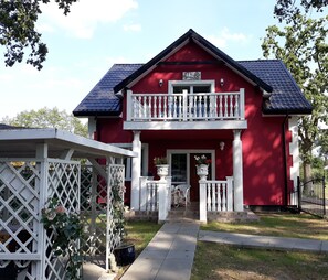 A red two-story house with a balcony, a white fence, and a pathway leading to the entrance.