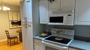 Una cocina con armarios blancos, microondas, horno y comedor con una silla y mesa.