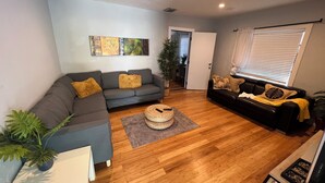 Una sala de estar moderna con un sofá seccional gris, un sofá negro y piso de madera. Hay un ventanal con persianas, una puerta que da a otra habitación y una obra de arte colgada en la pared.