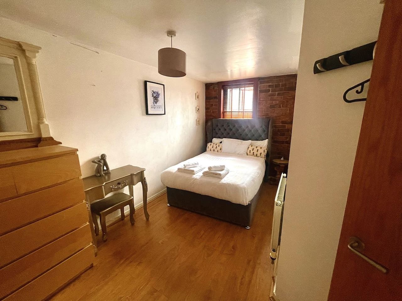 A bedroom with a bed, a desk, a chair, a dresser, a hanging light fixture, and a brick wall.