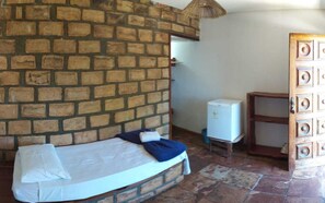 A room with a brick wall, a bed with a white mattress and blue blanket, a small white refrigerator, and a wooden cabinet with drawers. - Pousada Bosque do Lapão (Lençóis)
