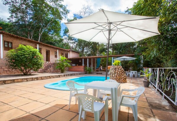A swimming pool with a white umbrella, surrounded by white plastic chairs and tables, with a stone wall and greenery in the background. - Pousada Bosque do Lapão (Lençóis)