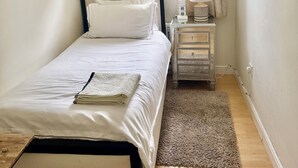 A neatly made bed with a white comforter and pillows, a bedside table with a lamp and a small plant, a window with curtains, and a wooden floor. - Sunny City (Peterborough)