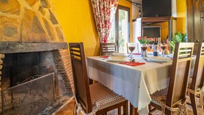 Un coin repas avec une cheminée en pierre, des chaises en bois, une table dressée pour deux personnes avec des verres à vin et des assiettes, et un téléviseur placé au-dessus de la cheminée.