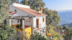 Un petit bâtiment blanc, surmonté d’un toit de tuiles rouges, entouré d’une végétation luxuriante et avec des montagnes en arrière-plan.