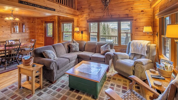 A cozy log cabin interior with a sectional sofa, a green coffee table, and a dining area with wooden chairs and a table. - Cozy mountain cabin near Sugarloaf—your four‑season adventure escape (Eustis)