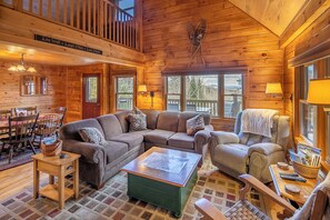 A cozy log cabin interior with a sectional sofa, a green coffee table, and a dining area with wooden chairs and a table. - Cozy mountain cabin near Sugarloaf—your four‑season adventure escape (Eustis)
