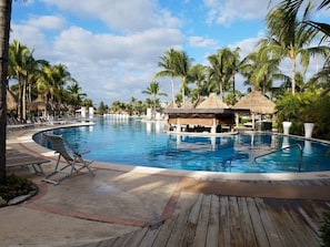 A resort pool with lounge chairs, palm trees, and thatched-roof structures.