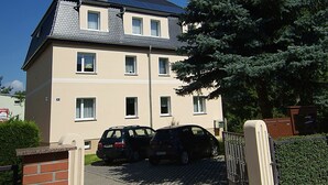 A multi-story residential building with a tiled roof, solar panels, and a gated driveway with two cars parked.