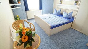 A bedroom with a single bed, a chair, a small table with a flower arrangement, a television on a shelf, and a window with curtains.