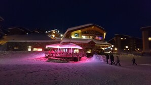 Zona exterior cubierta de nieve con un edificio de madera, un telesilla y un área de descanso cubierta con un toldo rojo.