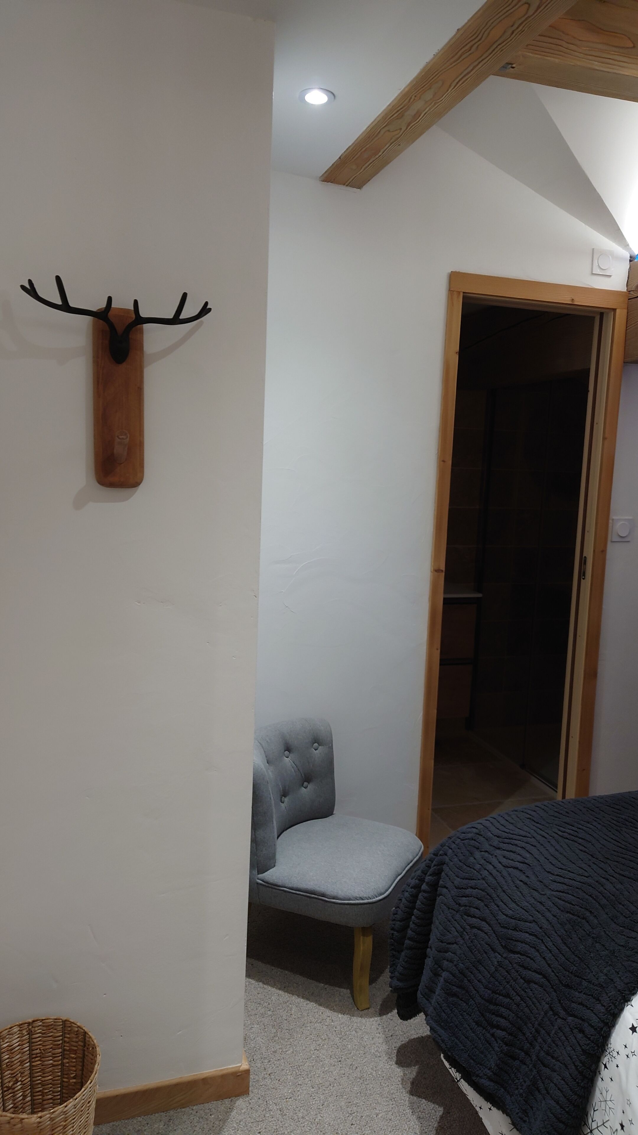 Une pièce avec un plafond en bois, une tête de cerf accrochée au mur, un fauteuil recouvert d'un tissu gris, une porte en bois et un panier en osier.