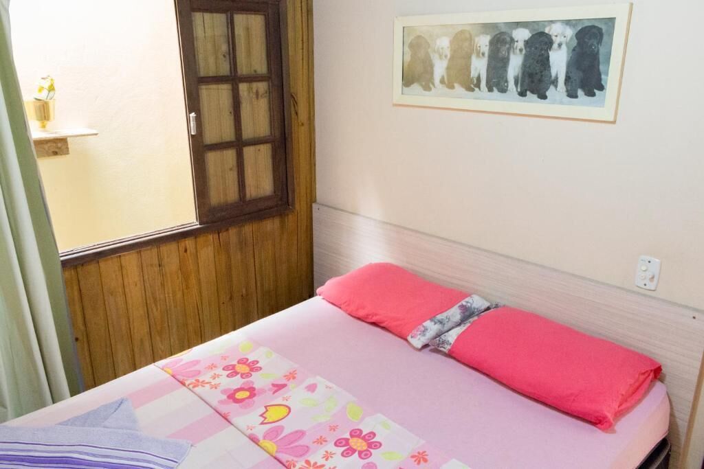 Un dormitorio con una cama, una ventana con persianas de madera, un cuadro colgado en la pared con perros y una pequeña estantería con un florero.