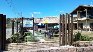 A fenced outdoor area with picnic tables and umbrellas, a reception sign, and a building with a sloped roof. - Pousada Vento & Cia (Laguna)