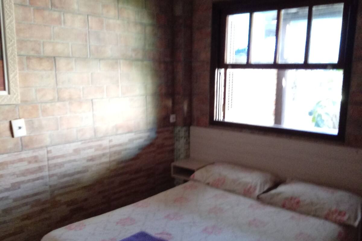 Un dormitorio con pared de ladrillo, ventana con persianas, cama con ropa de cama floral y un equipo de aire acondicionado.