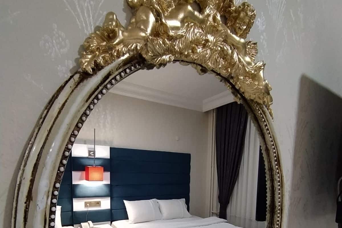 A hotel room with a large ornate mirror, two beds, a blue headboard, a bedside table with a lamp, and a nightstand with a phone.