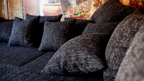 Ein dunkelgraues Sofa mit mehreren Kissen in einem Raum mit rustikalem Dekor, darunter eine Holzwand und ein Blumentopf.