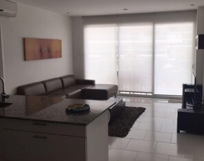 A modern living room with a sectional sofa, a kitchen island, and a large sliding glass door to the outdoors.